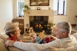 Elder couple sitting in front of a fire drinking hot chocolate from mugs.