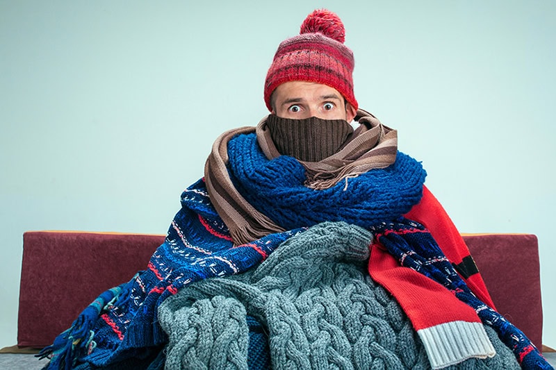 Man sitting on his couch bundled in many layers of jackets, scarfs, and blankets.