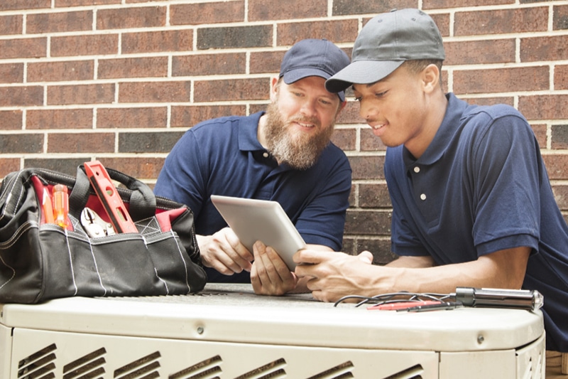 Two air conditioner repairmen working on a homes HVAC unit and using a digital tablet.